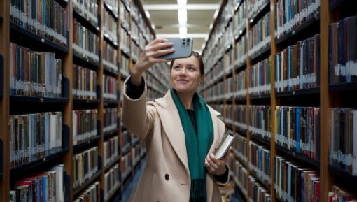 A woman in the stacks of a library taking a selfie with her phone.