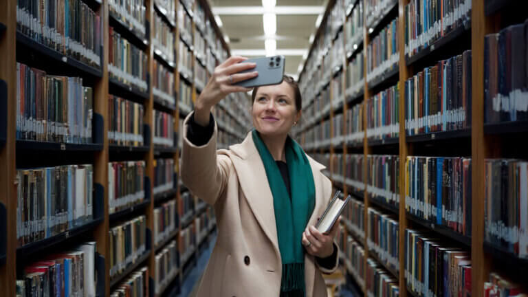 A woman in the stacks of a library taking a selfie with her phone.