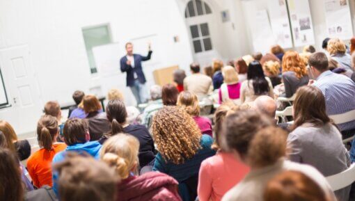 An audience in a lecture hall listening to a presenter at the front of the room.