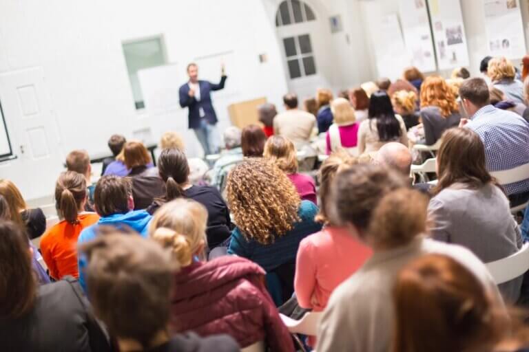 An audience in a lecture hall listening to a presenter at the front of the room.
