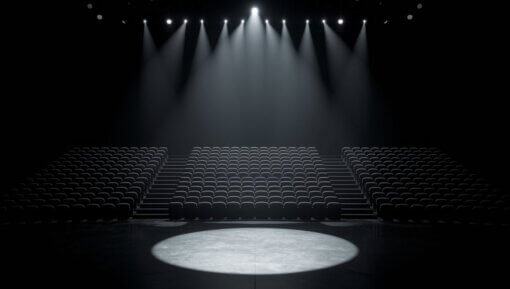 An empty stage, lit by a spotlight, with dark, empty seats in the background,
